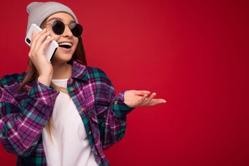 Closeup photo of beautiful happy positive young blonde woman wearing hipster purple shirt and casual white t-shirt grey hat and sunglasses isolated over red background holding in hand and