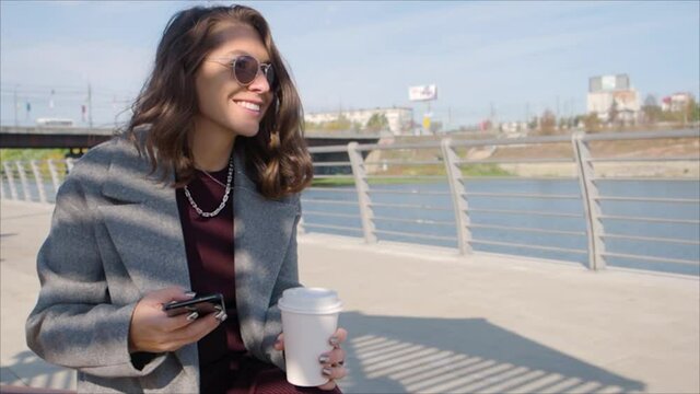 Woman with a cup of coffee typing text message on her smartphone outdoor while sitting on the city river coast.