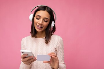 Photo of beautiful positive young brunette woman wearing pink casual sweater isolated over pink background wall wearing white bluetooth wireless headphones and listening to music and using mobile