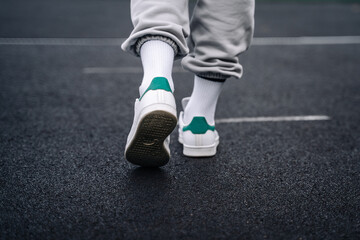 Fototapeta na wymiar Women's legs in sweatpants, white socks and white sneakers with green elements against the background of the track with markings in the stadium.