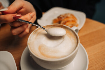 A woman's hand with a manicure holds a spoon while lifting the coffee foam in a cafe, on the background of pastries.