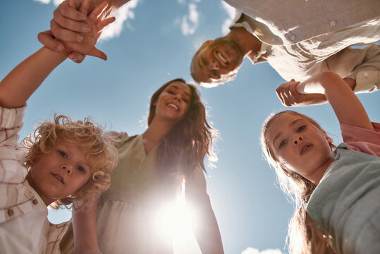 Portrait Of Lovely Young Family With Two Little Kids Holding Hands Together And Looking Down At Camera With The Blue Sky Above Them