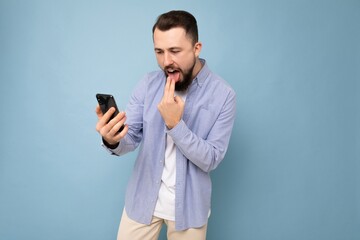 Photo shot of angry handsome bearded good looking young man wearing casual stylish outfit poising isolated on background with empty space holding in hand and using mobile phone messaging sms looking