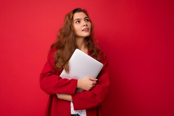 Thoughtful Beautiful smiling happy young brunet woman holding close computer laptop having an idea wearing red cardigan and white blouse looking at camera isolated over red background