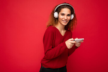 Pretty happy smiling young brunette curly woman wearing dark red sweater isolated on red backdrop wearing white headphones listening to music and using smartphone looking at camera