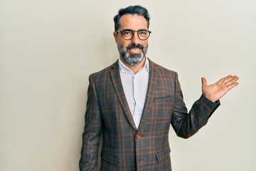 Middle age man with beard and grey hair wearing business jacket and glasses smiling cheerful presenting and pointing with palm of hand looking at the camera.