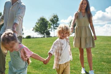 Cute little boy smiling at camera, holding hands together with his parents and sister and walking on green grass field in the park on a summer day