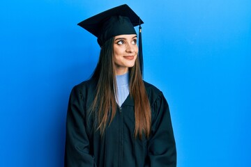 Beautiful brunette young woman wearing graduation cap and ceremony robe smiling looking to the side...