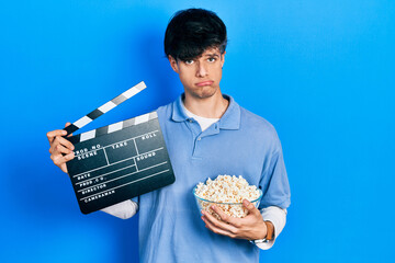 Handsome hipster young man eating popcorn holding cinema clapboard depressed and worry for...