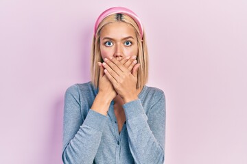 Beautiful blonde woman standing over pink background shocked covering mouth with hands for mistake....