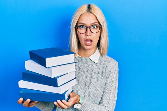 Beautiful blonde woman wearing glasses and holding pile of books in shock face, looking skeptical and sarcastic, surprised with open mouth
