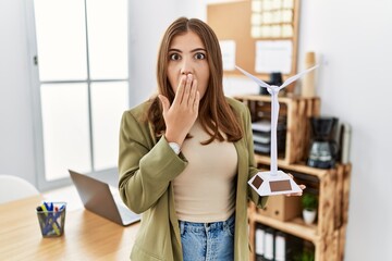 Young brunette woman holding solar windmill for renewable electricity at the office covering mouth...