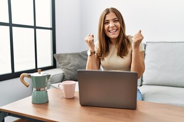 Young brunette woman using laptop at home drinking a cup of coffee celebrating surprised and amazed for success with arms raised and open eyes. winner concept.