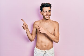 Young hispanic man wearing swimwear shirtless smiling and looking at the camera pointing with two...