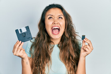 Young hispanic girl holding floppy disk and sdxc card angry and mad screaming frustrated and...