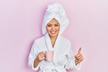 Young hispanic girl wearing bathrobe drinking cup of coffee smiling with an idea or question pointing finger with happy face, number one