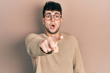 Young hispanic man wearing casual clothes and glasses pointing with finger surprised ahead, open mouth amazed expression, something on the front