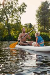 Father teaching his child, little daughter sup surfing on a river surrounded by the beautiful...