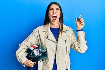 Young hispanic woman holding motorcycle helmet and key angry and mad screaming frustrated and...