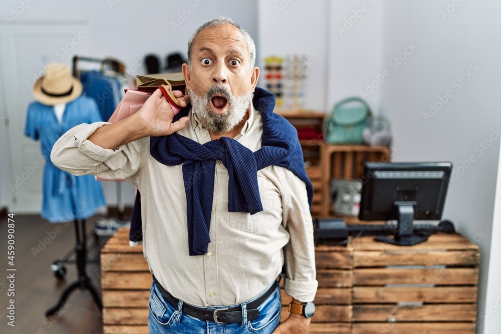 Wall mural Handsome senior man holding shopping bags at boutique shop in shock face, looking skeptical and sarcastic, surprised with open mouth