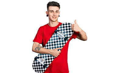 Young caucasian boy with ears dilation holding skate smiling happy and positive, thumb up doing excellent and approval sign
