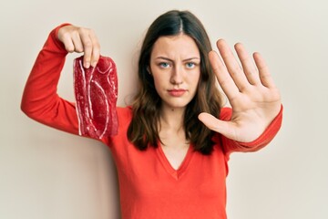 Young brunette woman holding raw beef steak with open hand doing stop sign with serious and...