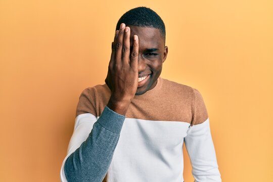 Young african american man wearing casual clothes covering one eye with hand, confident smile on face and surprise emotion.