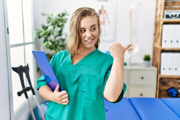 Young caucasian woman working at pain recovery clinic smiling with happy face looking and pointing to the side with thumb up.