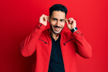 Young hispanic man wearing red leather jacket smiling pulling ears with fingers, funny gesture....