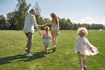 Fototapeta premium Happy parents holding hands, playing on grass field with their little kids, boy and girl, having fun together on a summer day