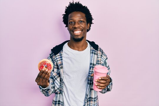 Young african american man eating doughnut and drinking coffee smiling with a happy and cool smile on face. showing teeth.