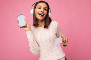 Naklejka premium Beautiful happy smiling young woman wearing stylish casual outfit isolated on background wall holding and showing mobile phone with empty display for mockup and credit card wearing white bluetooth
