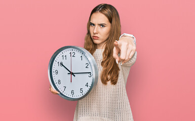 Young blonde woman holding big clock pointing with finger to the camera and to you, confident gesture looking serious