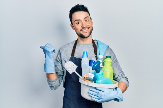 Young hispanic man holding cleaning products pointing thumb up to the side smiling happy with open mouth