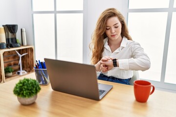 Young caucasian woman working at the office using computer laptop checking the time on wrist watch, relaxed and confident