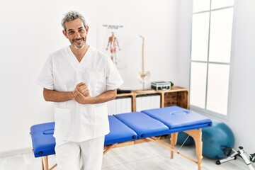 Fototapeta na wymiar Middle age hispanic therapist man working at pain recovery clinic with hands together and crossed fingers smiling relaxed and cheerful. success and optimistic