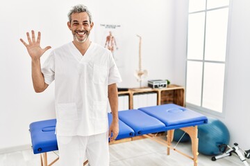Obraz premium Middle age hispanic therapist man working at pain recovery clinic showing and pointing up with fingers number five while smiling confident and happy.