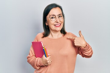 Middle age hispanic woman holding book and color pencils pointing finger to one self smiling happy...