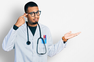 Young african american man wearing doctor uniform and stethoscope confused and annoyed with open palm showing copy space and pointing finger to forehead. think about it.