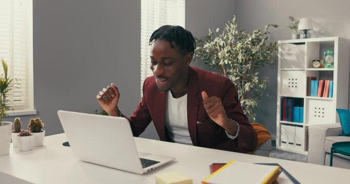 Smiling happy employee, boss of the company, wins tender, contract, earned big money, raises hands in gesture of victory, winning, achieved success, dark-skinned man in jacket sits in front of laptop