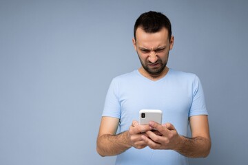 Photo of dissatisfied handsome young man with beard wearing everyday blue t-shirt isolated over blue background holding and using mobile phone communication online on the internet looking at gadjet