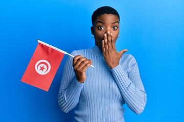Young african american woman holding tunisia flag covering mouth with hand, shocked and afraid for...