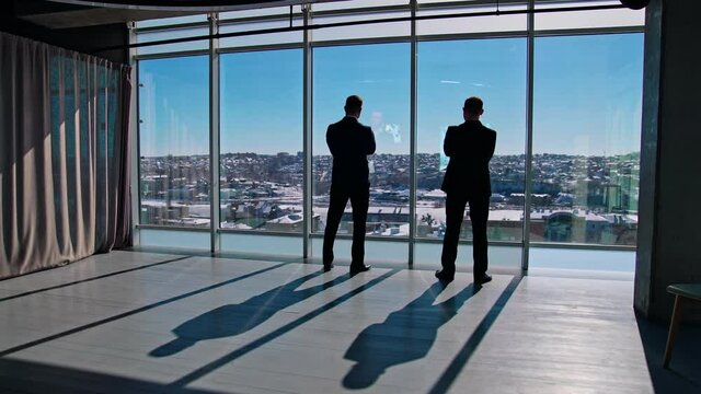 Business people talk while looking from office windows. Male business partners standing near large window indoors at sunny day. Rear view.