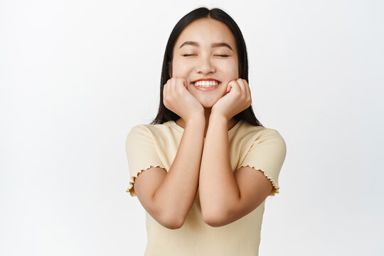 Happy asian woman dreaming, smiling with closed eyes, imaging something romantic, standing over white background