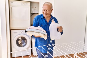 Senior caucasian man holding detergent and folded clothes standing by washing machine at the...