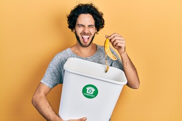 Handsome hispanic man recycling banana peel composting organic fertilizer sticking tongue out happy...