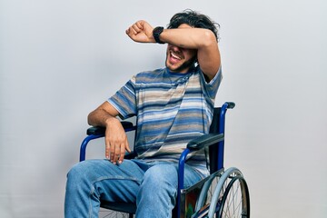 Handsome hispanic man sitting on wheelchair covering eyes with arm smiling cheerful and funny. blind concept.