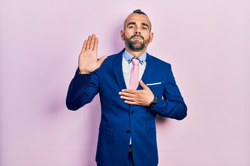 Young hispanic man wearing business suit and tie swearing with hand on chest and open palm, making...