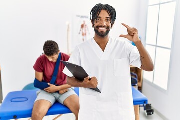 Obraz premium Young hispanic man working at pain recovery clinic with a man with broken arm smiling and confident gesturing with hand doing small size sign with fingers looking and the camera. measure concept.