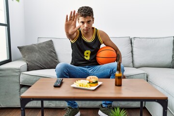 Young handsome hispanic man holding basketball ball cheering tv game with open hand doing stop sign with serious and confident expression, defense gesture